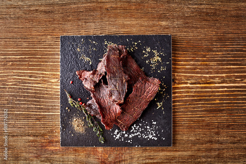 Картината върху платно Dried peppered beef jerky cut in strips on the black board on wooden background