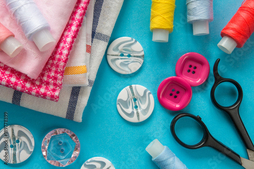 Multicolored threads, scissors, buttons, fabric and various sewing accessories on a blue background with copy space flat lay