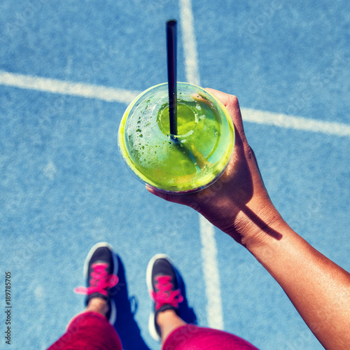 Green smoothie woman drinking plastic cup breakfast meal takeaway to go after morning run on blue tracks. Healthy lifestyle sporty person pov of hand holding glass with running shoes feet selfie.