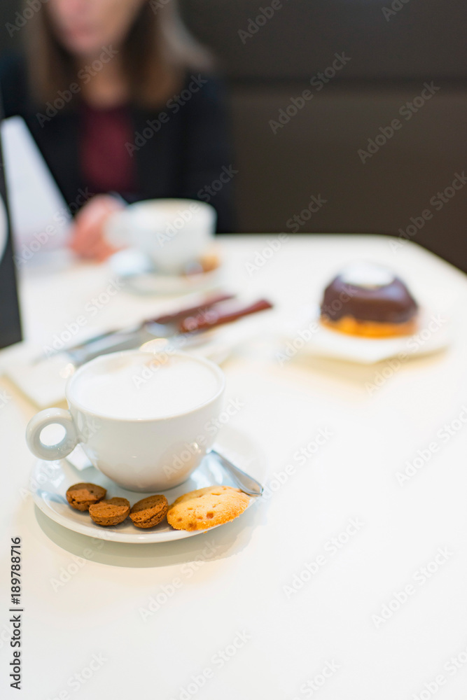 Cappuccino with cookies on white table with woman blurred in background.