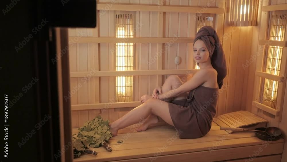 Young woman relaxing in a sauna dressed in a towel. Interior of new Finnish sauna, infrared panels for medical procedures, classic wooden sauna. Young woman relaxing in a sauna dressed in a towel. Interior of new Finnish sauna, infrared panels for medical procedures, classic wooden sauna.
