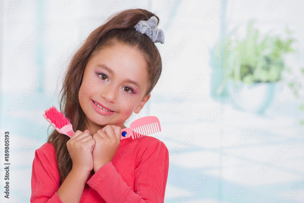 Little beautiful girl wearing a red blouse and holding a hair brush and comb in her hand, in a blurred background Little beautiful girl wearing a red blouse and holding a hair brush and comb in her hand, in a blurred background