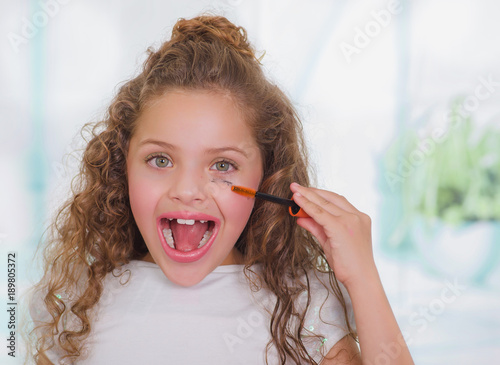 Close up of surprised little girl using wrong an eye mascara in her face in a blurred background