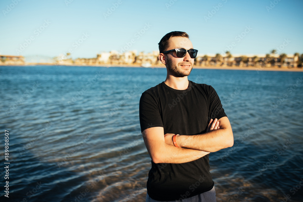 Closeup of attractive young man in sunglasses in black t-shirt standing on the sea beach