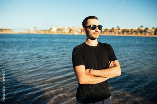 Closeup of attractive young man in sunglasses in black t-shirt standing on the sea beach