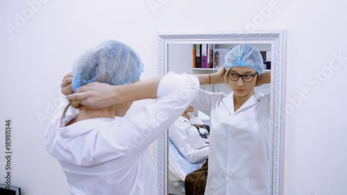 cosmetology room in the clinic, a female doctor wearing glasses, wearing a white uniform, dresses a medical cap and bandage. stands in front of a mirror. photo