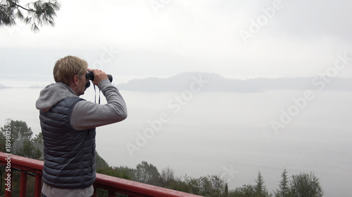 Man with binoculars looking out over misty nature landscape.