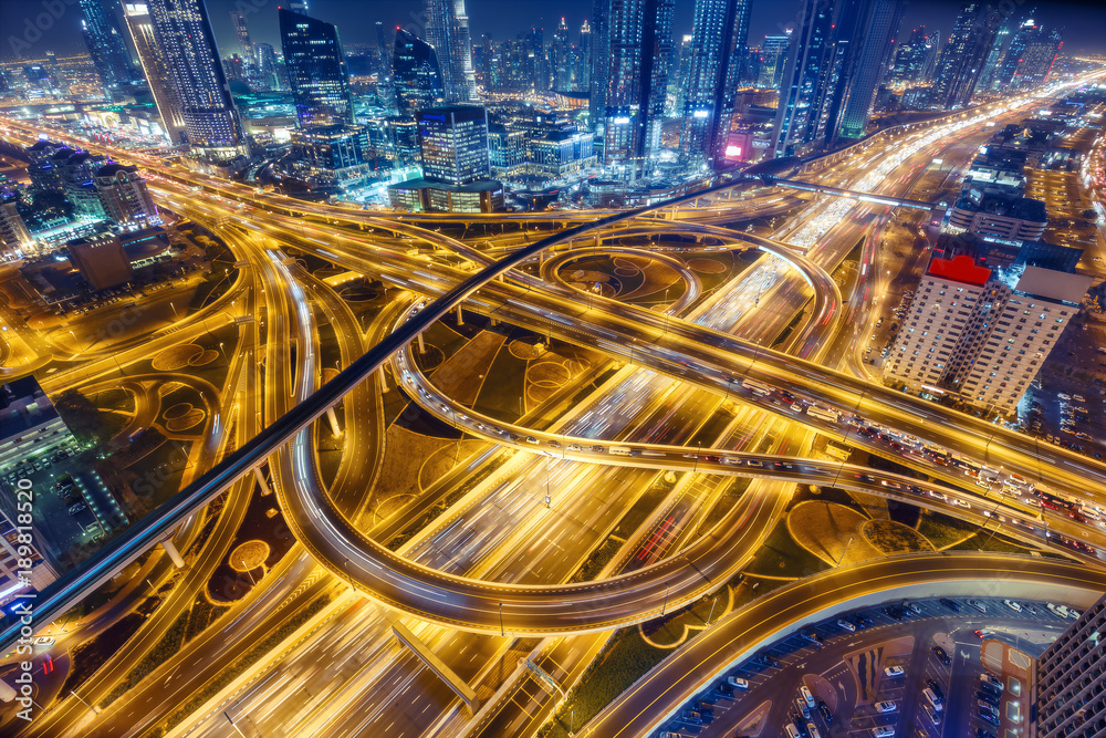 Naklejka premium Widok z lotu ptaka na duże skrzyżowanie autostrad z ruchem w Dubaju, Zjednoczone Emiraty Arabskie, w nocy. Malowniczy pejzaż. Kolorowe tło transportu, komunikacji i jazdy.