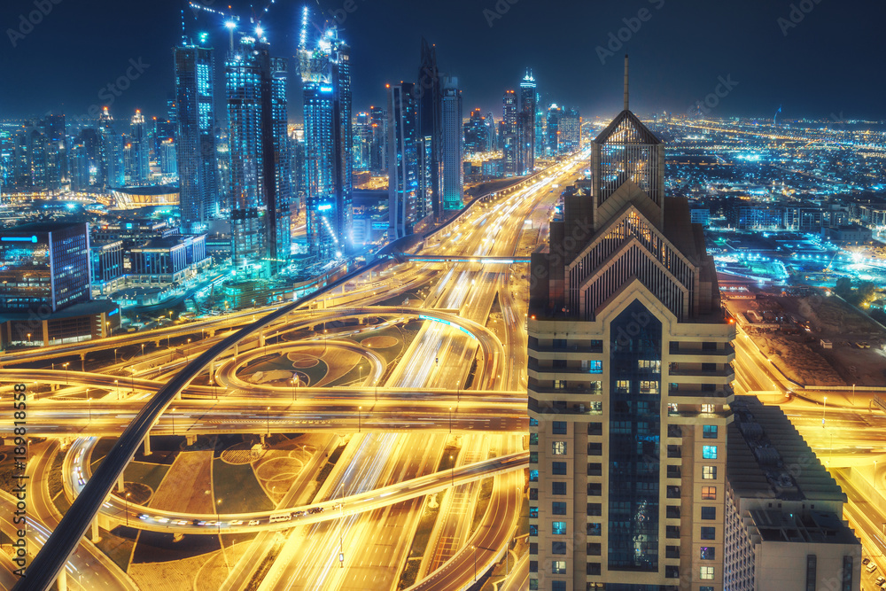 Naklejka premium Aerial view of big highway interchange with traffic in Dubai, UAE, at night. Scenic cityscape. Colorful transportation, communications and driving background.