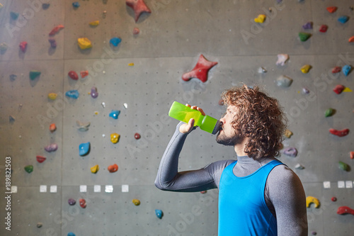 Active guy drinking water from plastic bottle on background of climbing wall