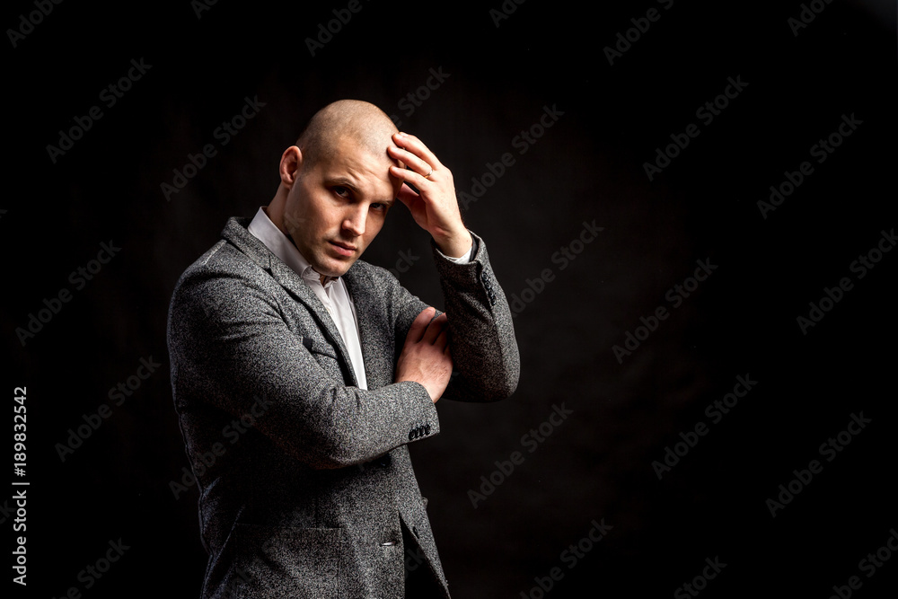 A young bald man in a white shirt and gray suit is upset by a serious problem in business, at work on a black isolated background. A man has a headache