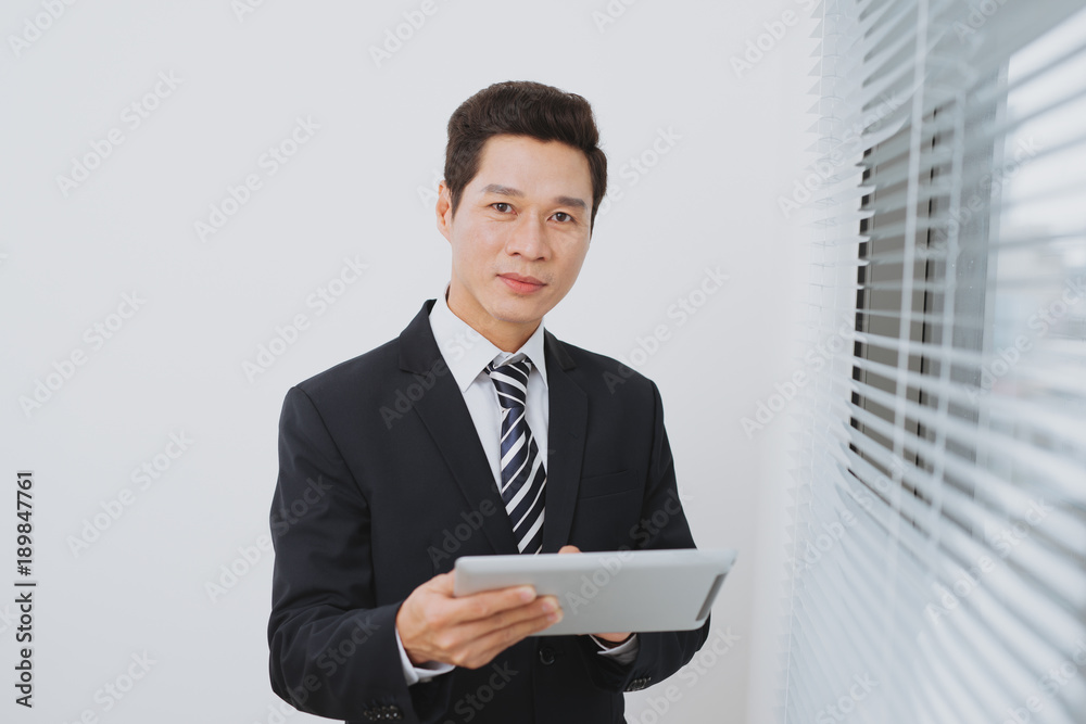 Smiling asian businesman holding digital tablet standing by the window. Smiling asian businesman holding digital tablet standing by the window.