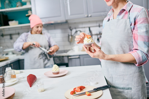 Food stylist putting piece of fresh strawberry on top of cupcake decorated by whipped cream