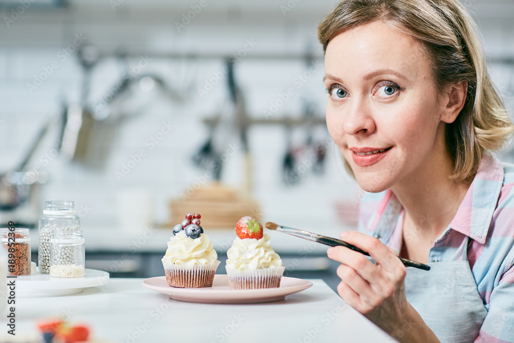 Pretty young woman with paintbrush looking at camera in process of decorating muffin tops Pretty young woman with paintbrush looking at camera in process of decorating muffin tops