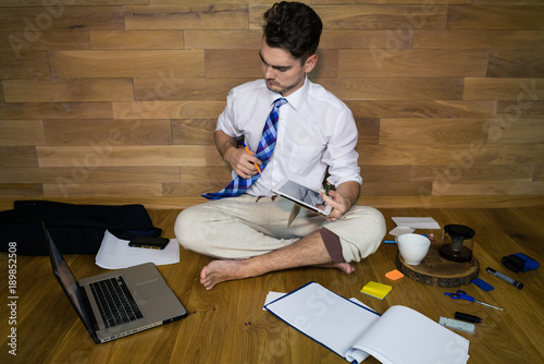 A barefoot businessman sits on the floor against a wall and using a laptop for read important news. Remote working, thinking about a solution. Young man working at home in funny clothes.
