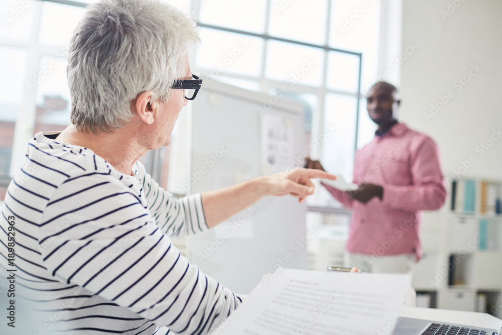 Mature grey-haired man pointing aside while commenting report of colleague by whiteboard at start-up meeting