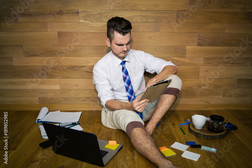 Serious young man works with project in tablet. A barefoot businessman in funny clothes sits on the floor against a wooden wall with laptop and other gadgets