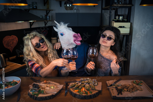 Two beautiful women with funny unicorn has dinner together. Group of young people drinks wine and enjoying evening. Man in a funny rubber mask. Crazy party in modern home kitchen at the bar counter