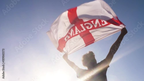 Young man holding england national flag to the sky with two hands at the beach at sunset uk united kingdom