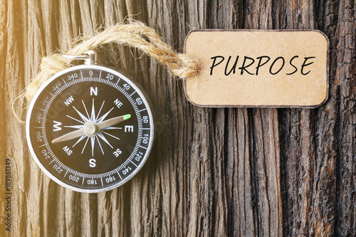 Magnetic compass and paper tag written with PURPOSE on old wooden background.