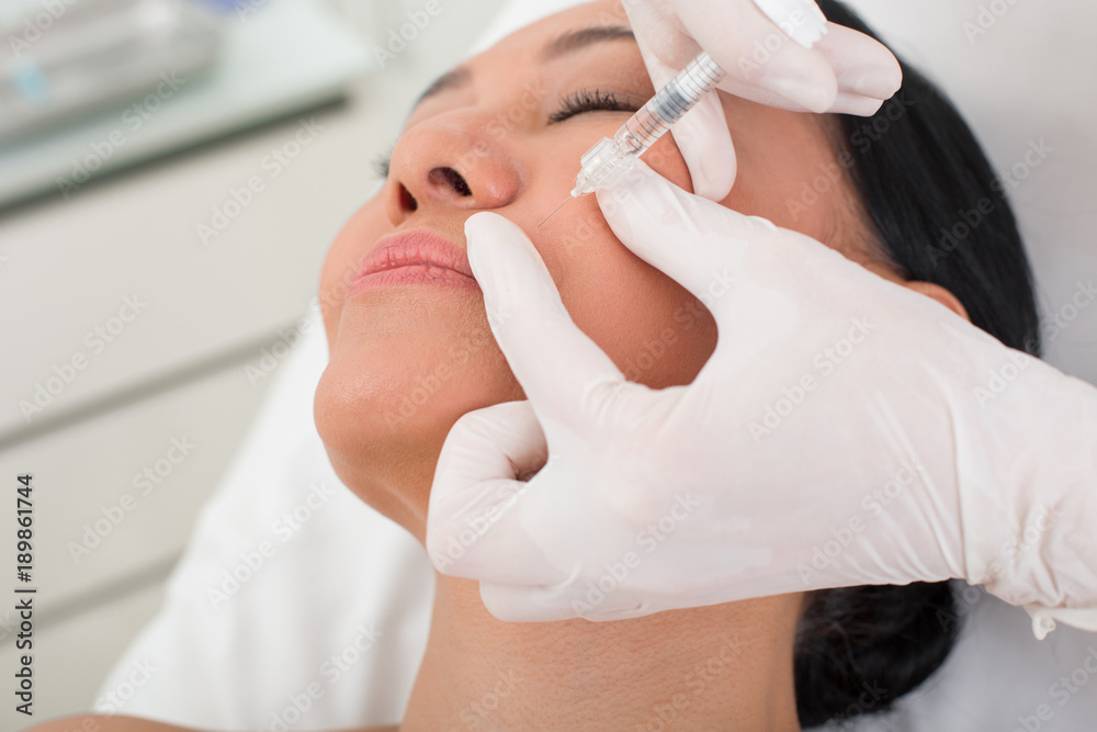 Fototapeta premium Close up of female face. Tranquil middle-aged lady lying on the couch with closed eyes. Therapist taking care of skin by botox injection of patient at spa