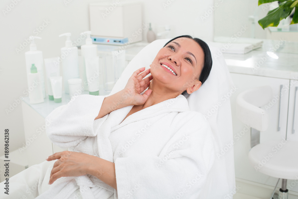 Happy asian woman lying on the couch at spa. She is touching her neck with pleased smile and wearing bathrobe Happy asian woman lying on the couch at spa. She is touching her neck with pleased smile and wearing bathrobe