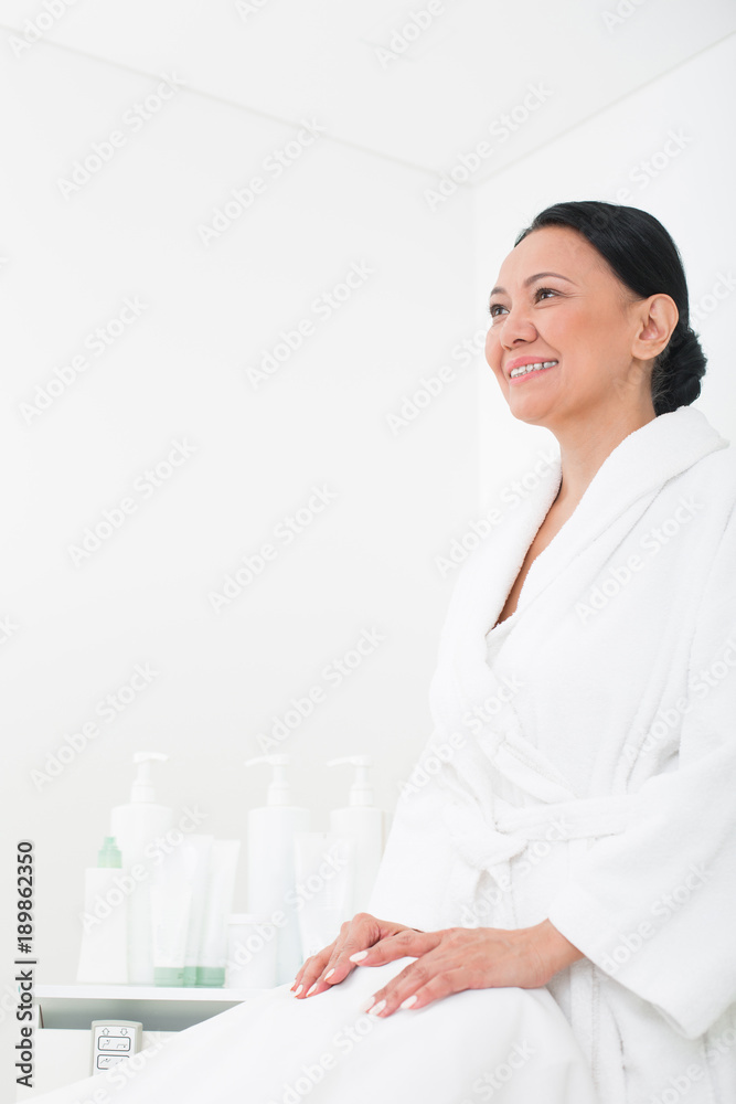 Pleased asian woman smiling and sitting in wellness center. She is wearing bathrobe. Copy space in left side Pleased asian woman smiling and sitting in wellness center. She is wearing bathrobe. Copy space in left side