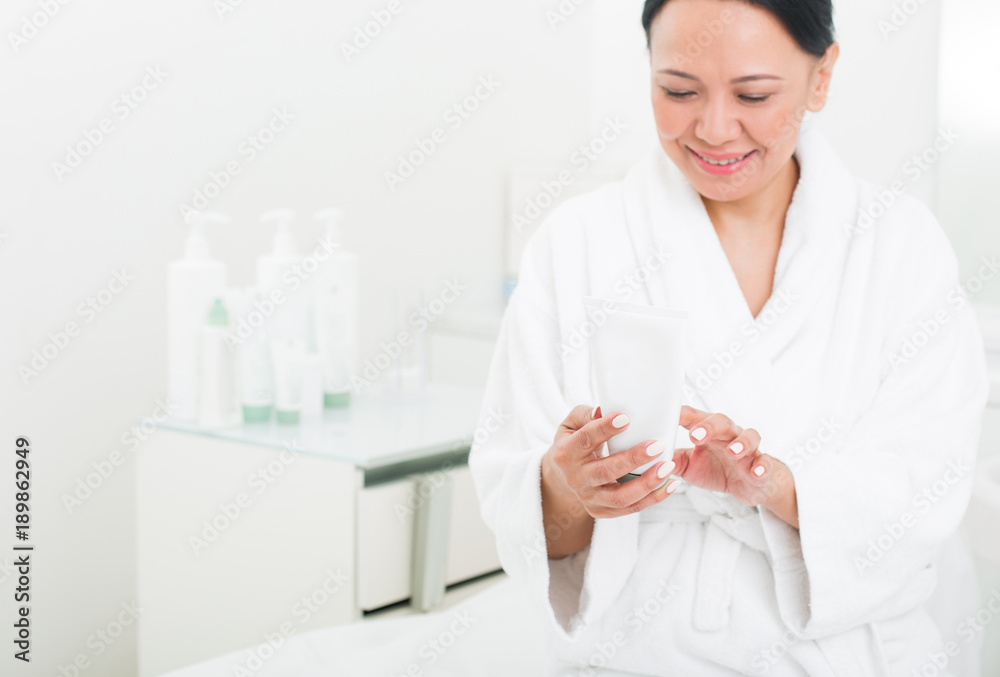 Happy senior lady smiling and holding a tube of cream in beauty salon. She is looking at packaging and reading the instruction. Copy space in left side Happy senior lady smiling and holding a tube of cream in beauty salon. She is looking at packaging and reading the instruction. Copy space in left side