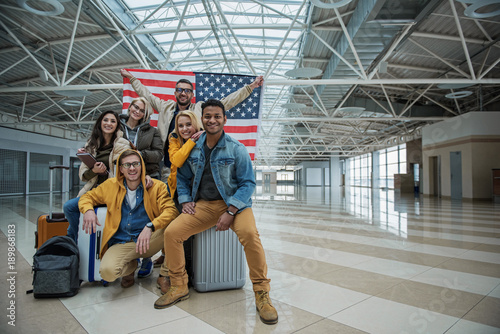 We love our country. Full length portrait of optimistic young friends are sitting on suitcases at airport with usa flag. They are looking at camera with smile while expressing happiness. Copy space