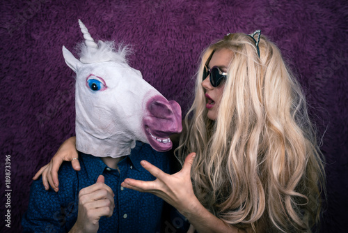 Portrait of happy unicorn in the arms of freaky young woman in sunglasses and kitty ears on the purple background. Expressive blond with strange guy. Unusual people shows emotions.