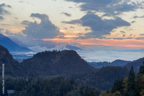 There are a lot of clouds in the distance as the sun rises. There are mountains and trees in the foreground and the sun rise in the background. Taken at Lake Bled in Slovenia.