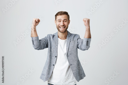 Happy lucky overjoyed male winner with stubble in trendy clothes clenches fists, rejoices triumph at work, glad to recieve high profits, has own prosperous business. Good-looking guy achieves goals