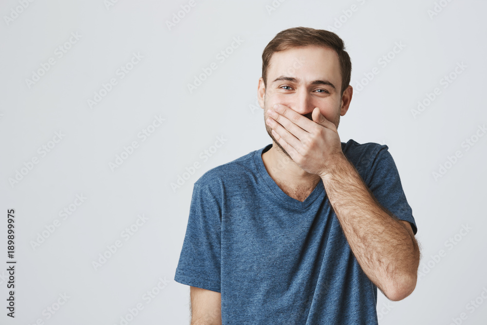 Positive human expressions concept. Bearded caucasian male in blue t-shirt laughs at good joke, hides his smile behind his hand. Laughing man posing against gray studio wall. Positive human expressions concept. Bearded caucasian male in blue t-shirt laughs at good joke, hides his smile behind his hand. Laughing man posing against gray studio wall.