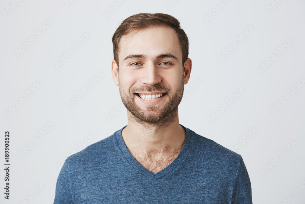 Cheerful handsome man with beard wearing blue t-shirt smiling happily while receiving positive news. Handsome caucasian male model looking at camera with joyful smile posing against studio wall. Cheerful handsome man with beard wearing blue t-shirt smiling happily while receiving positive news. Handsome caucasian male model looking at camera with joyful smile posing against studio wall.