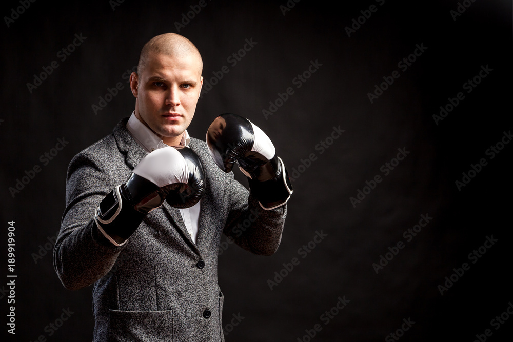 Plakat A young man manager in a white shirt, gray suit, boxing gloves looks seriously and boxing on a black isolated background