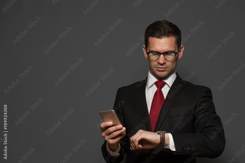 Portrait of successful young man hurrying on meeting and looking at his watch with smartphone in his hand on gray background. Copyspace Portrait of successful young man hurrying on meeting and looking at his watch with smartphone in his hand on gray background. Copyspace