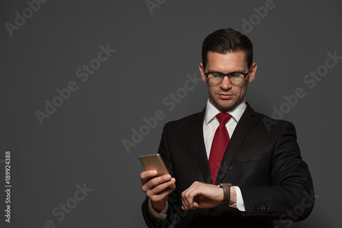 Portrait of successful young man hurrying on meeting  and looking at his watch with  smartphone in his hand on gray background. Copyspace © pavel_shishkin