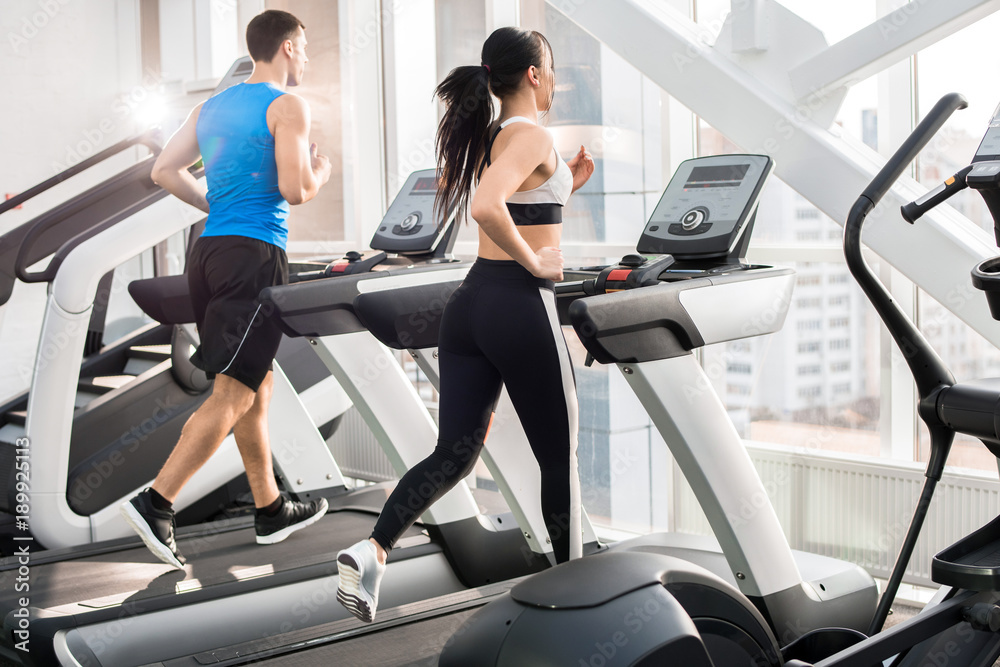 Back view of two fit young people, man and woman, running on treadmills facing windows in modern gym, copy space