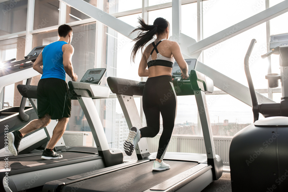Full length portrait of two fit young people, man and woman, running on treadmills facing windows in modern gym, copy space Full length portrait of two fit young people, man and woman, running on treadmills facing windows in modern gym, copy space