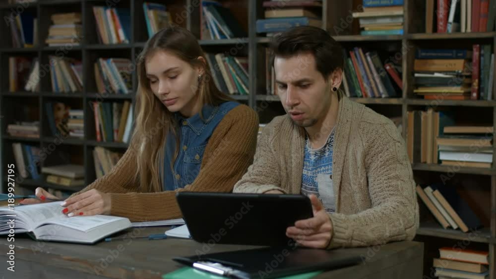 Smart couple of hipster students with digital tablet pc and books researching in campus library over bookshelf background. Two attractive c students studying together using tablet and books in library