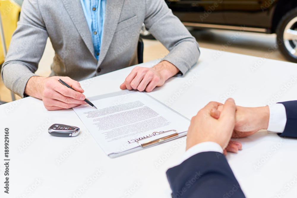 Close up of young handsome man reading purchase contract sitting at desk in dealership shop buying luxury car, copy space Close up of young handsome man reading purchase contract sitting at desk in dealership shop buying luxury car, copy space