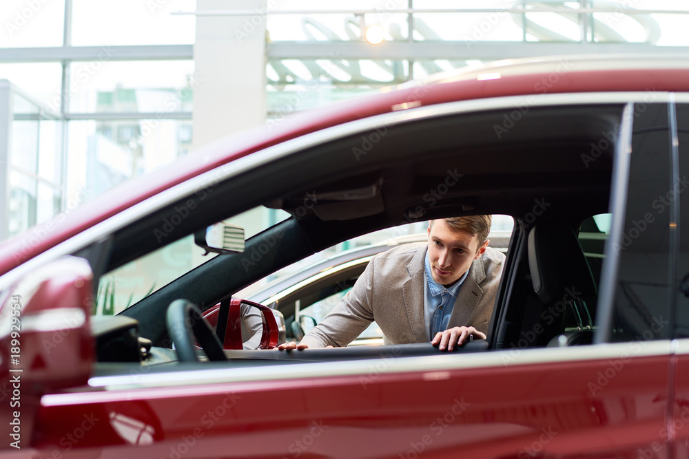 Portrait of handsome young man choosing new car in showroom looking inside open windows at car interior, copy space Portrait of handsome young man choosing new car in showroom looking inside open windows at car interior, copy space