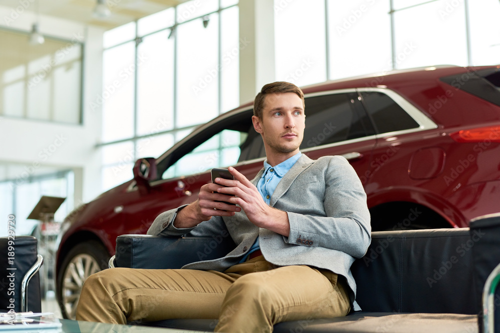 Portrait of handsome young man sitting on sofa in car showroom against background of shiny luxury cars, copy space