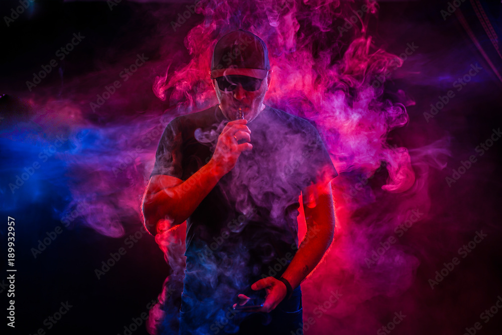 A man smokes an electronic cigarette. The man in the smoke. bearded man vaping. Men with beard in sunglasses vaping and releases a cloud of vapor. vaping man holding a mod. A cloud of vapor. 