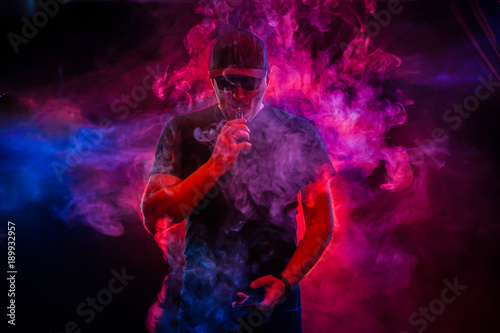 A man smokes an electronic cigarette. The man in the smoke. bearded man vaping. Men with beard in sunglasses vaping and releases a cloud of vapor. vaping man holding a mod. A cloud of vapor. 