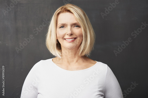 Confident blond female portrait. Close-up studio shot of a beautiful senior woman wearing shirt while standing at dark background and smiling.  © gzorgz