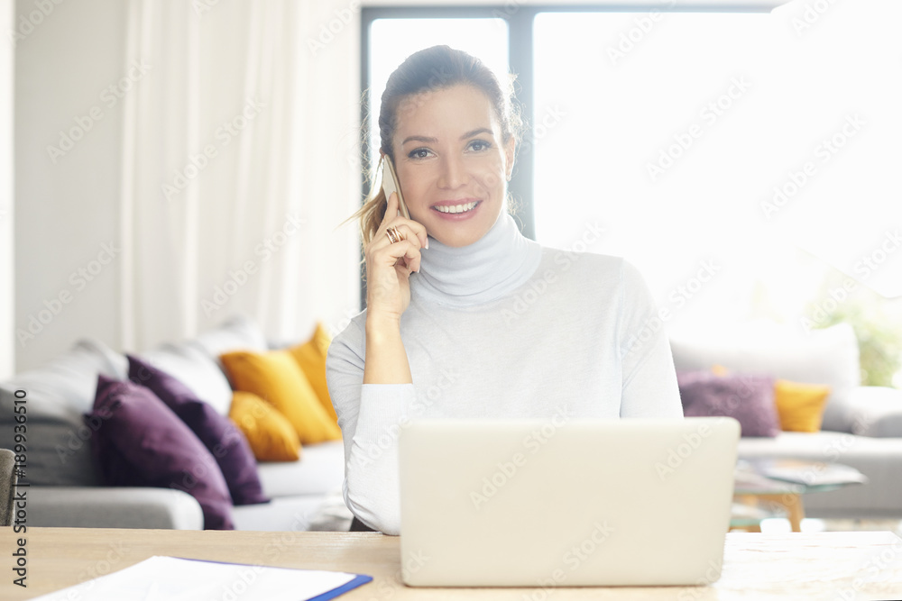 Home office. Confident woman talking on her mobile phone and using laptop while sitting at desk and working from home. Home office. Confident woman talking on her mobile phone and using laptop while sitting at desk and working from home.
