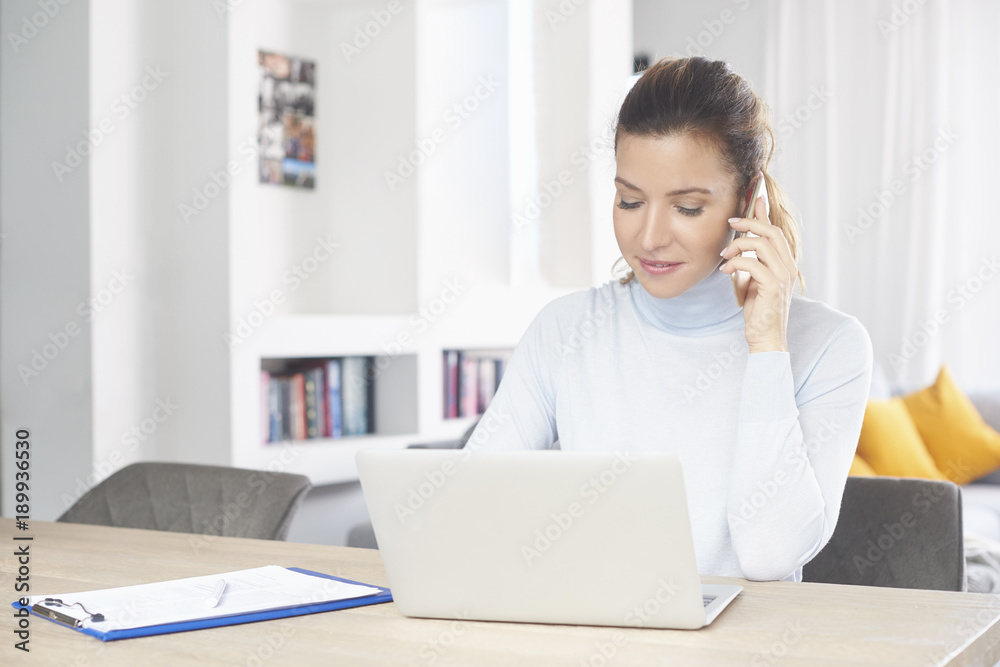 Home office. Confident woman talking on her mobile phone and using laptop while sitting at desk and working from home. Home office. Confident woman talking on her mobile phone and using laptop while sitting at desk and working from home.