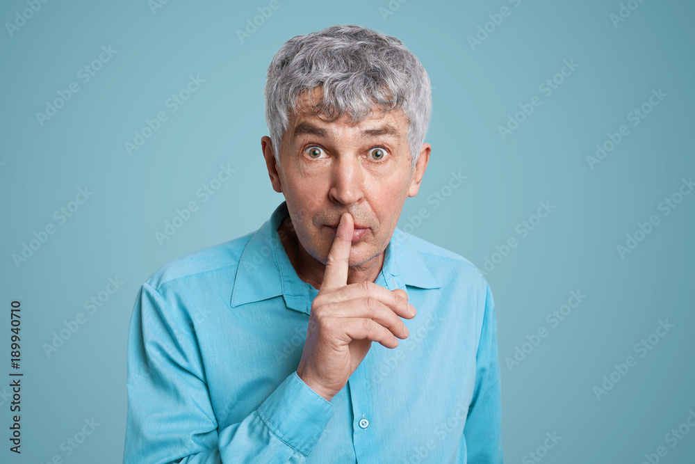 Horizontal shot of grey haired male looks with puzzled expression at camera, keeps fore finger on lips, asks to be quiet and mute, demands hush, dressed in elegant formal shirt isolated over blue wall Horizontal shot of grey haired male looks with puzzled expression at camera, keeps fore finger on lips, asks to be quiet and mute, demands hush, dressed in elegant formal shirt isolated over blue wall