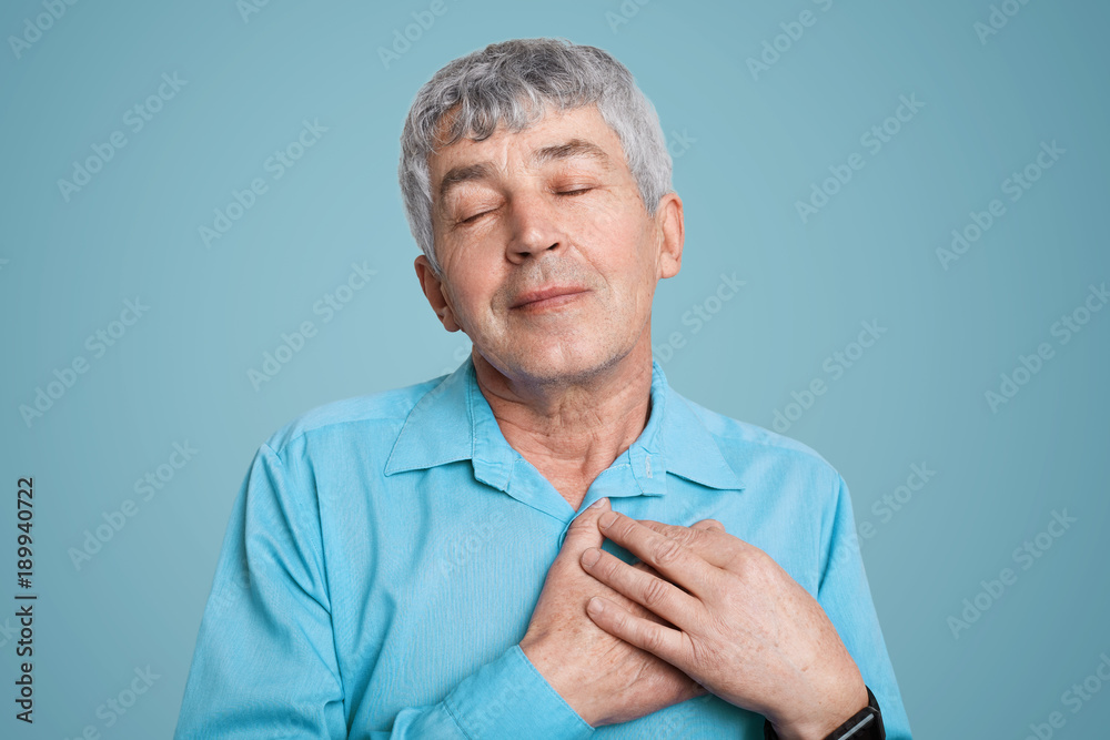Handsome pleased grey haired grandfather keeps hands on chest, expresses his great love to children, dress in formal wear as has special occasion today, isolated over blue studio background. Handsome pleased grey haired grandfather keeps hands on chest, expresses his great love to children, dress in formal wear as has special occasion today, isolated over blue studio background.